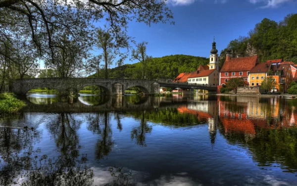 HD desktop wallpaper showcasing a serene man-made town with historic buildings, a stone bridge, and their reflections on calm river water under a clear blue sky.