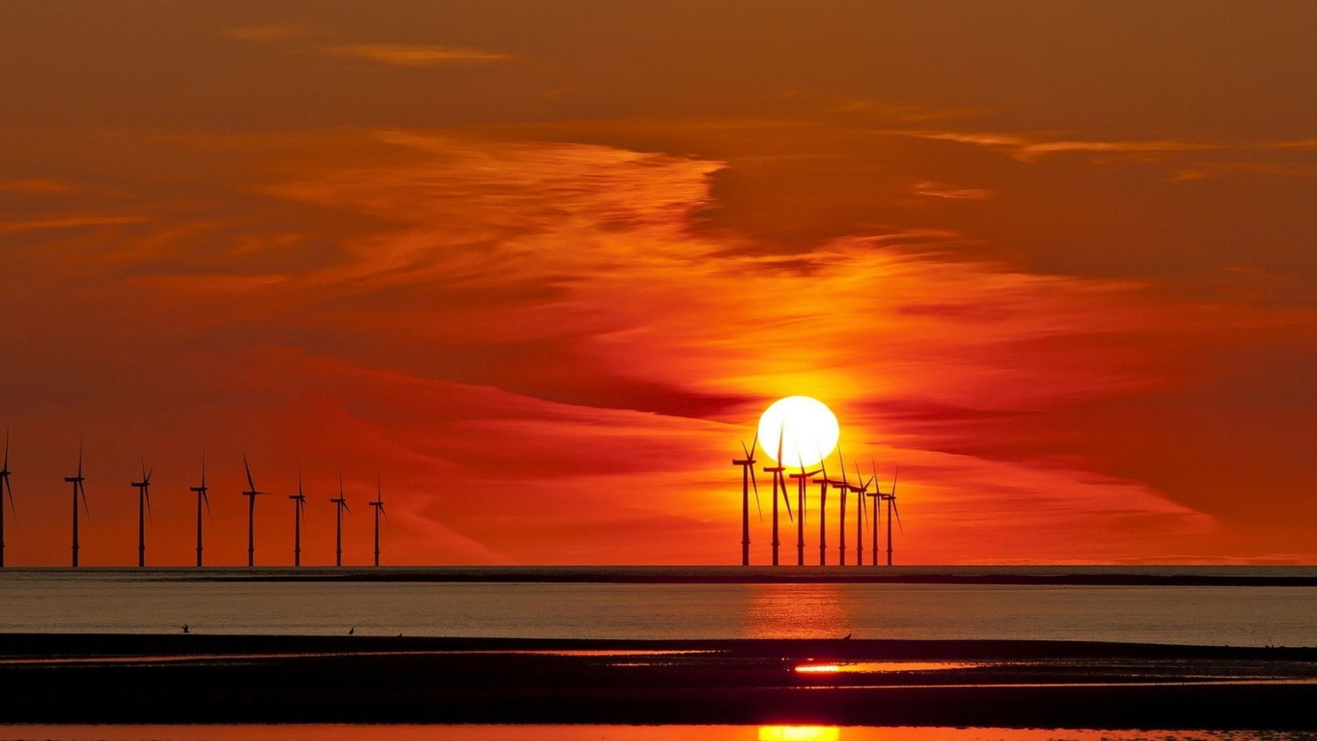 HD desktop wallpaper featuring a row of man-made windmills silhouetted against a vibrant orange sunset over calm water.