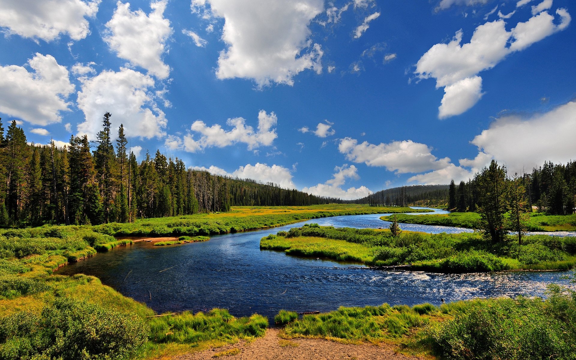 HD desktop wallpaper featuring a serene river winding through lush green meadows under a bright blue sky with scattered clouds in a vibrant natural landscape.