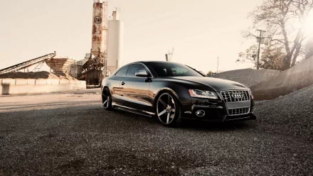 A sleek black Audi S5 sits prominently on a gravel surface, framed by industrial structures in the background, creating a striking HD desktop wallpaper.