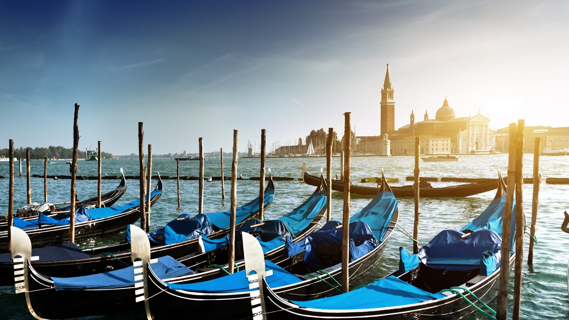 HD desktop wallpaper featuring man-made gondolas lined up on the water in Venice, with the city's historic architecture and a bright sky in the background.