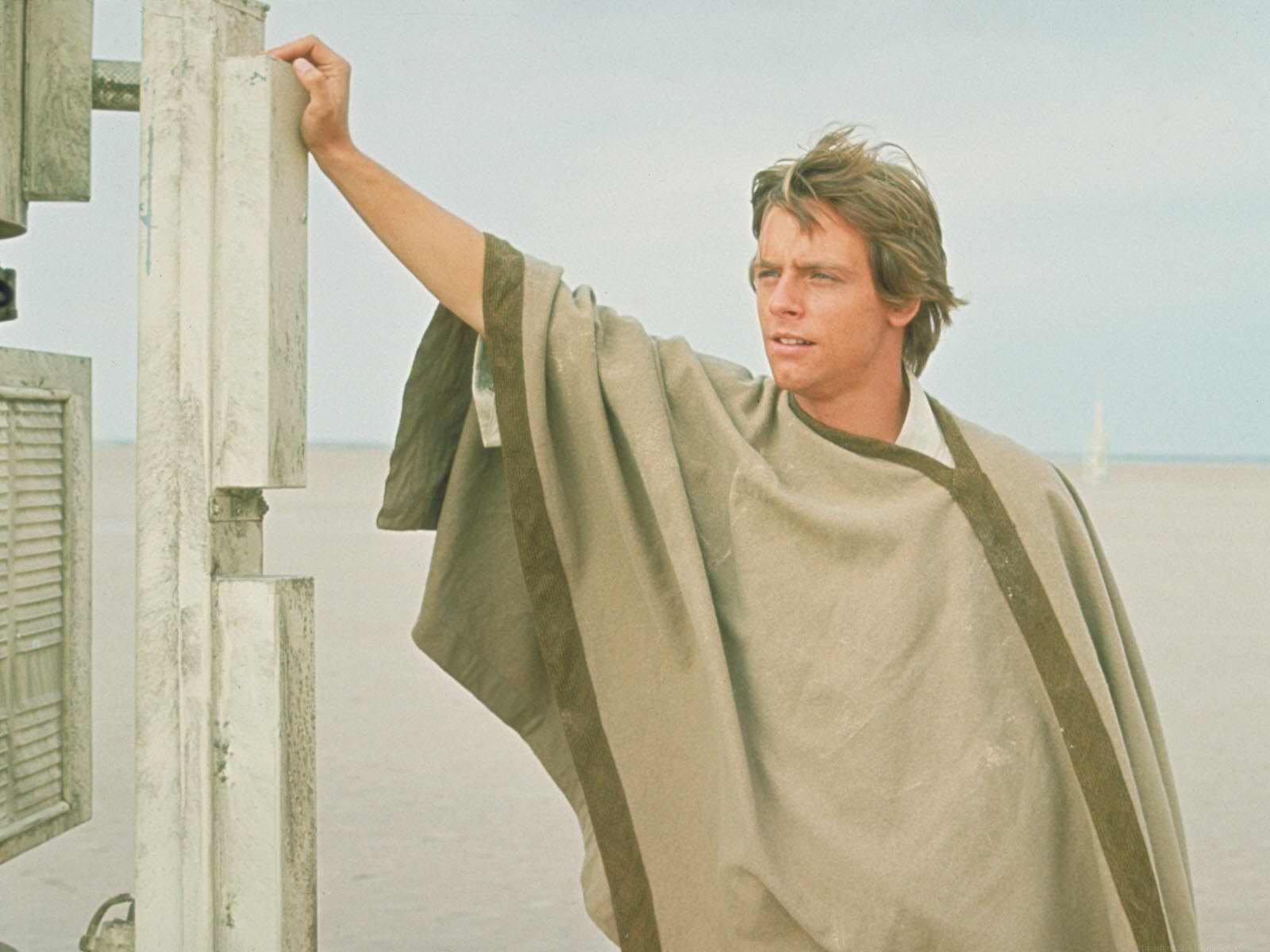HD PC desktop wallpaper: a robed young man from the movie Star Wars leans on a weathered post against a pale desert horizon.