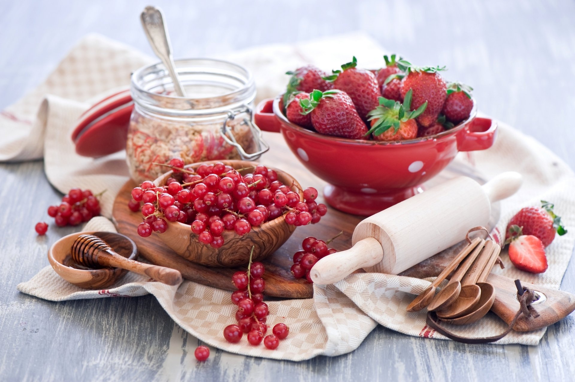 A vibrant still life featuring fresh strawberries and red currants arranged with a glass jar and wooden utensils, creating a colorful and inviting food scene. HD desktop wallpaper.