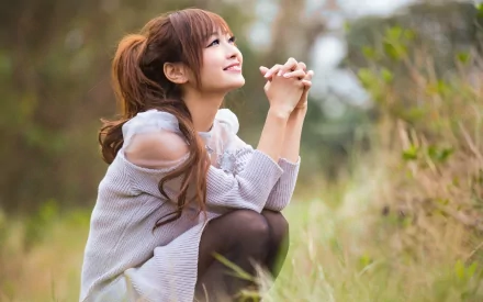 HD desktop wallpaper of an Asian woman kneeling in a grassy field, looking up with a smile, exuding a serene and hopeful atmosphere.