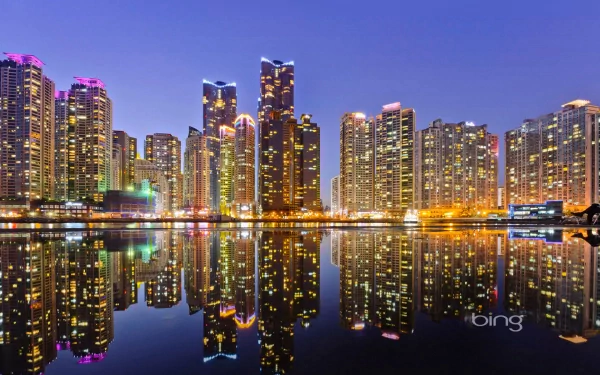 A vibrant cityscape of Busan at twilight, featuring towering skyscrapers illuminated in colorful lights, reflecting beautifully on the water's surface. A stunning HD desktop wallpaper.