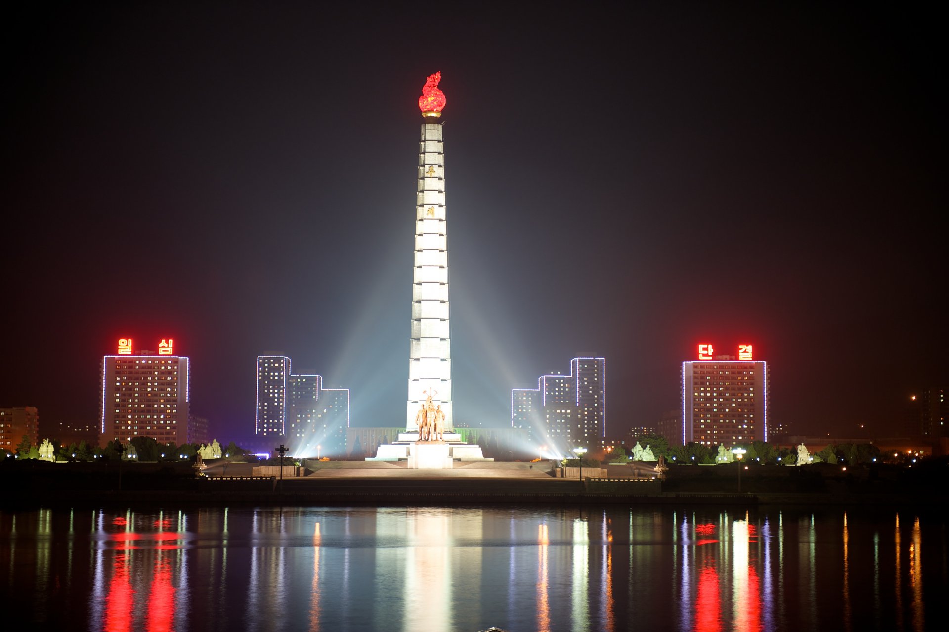 A nighttime 4K Ultra HD wallpaper featuring the illuminated Juche Tower, a man-made landmark, with city buildings and reflections on the water.