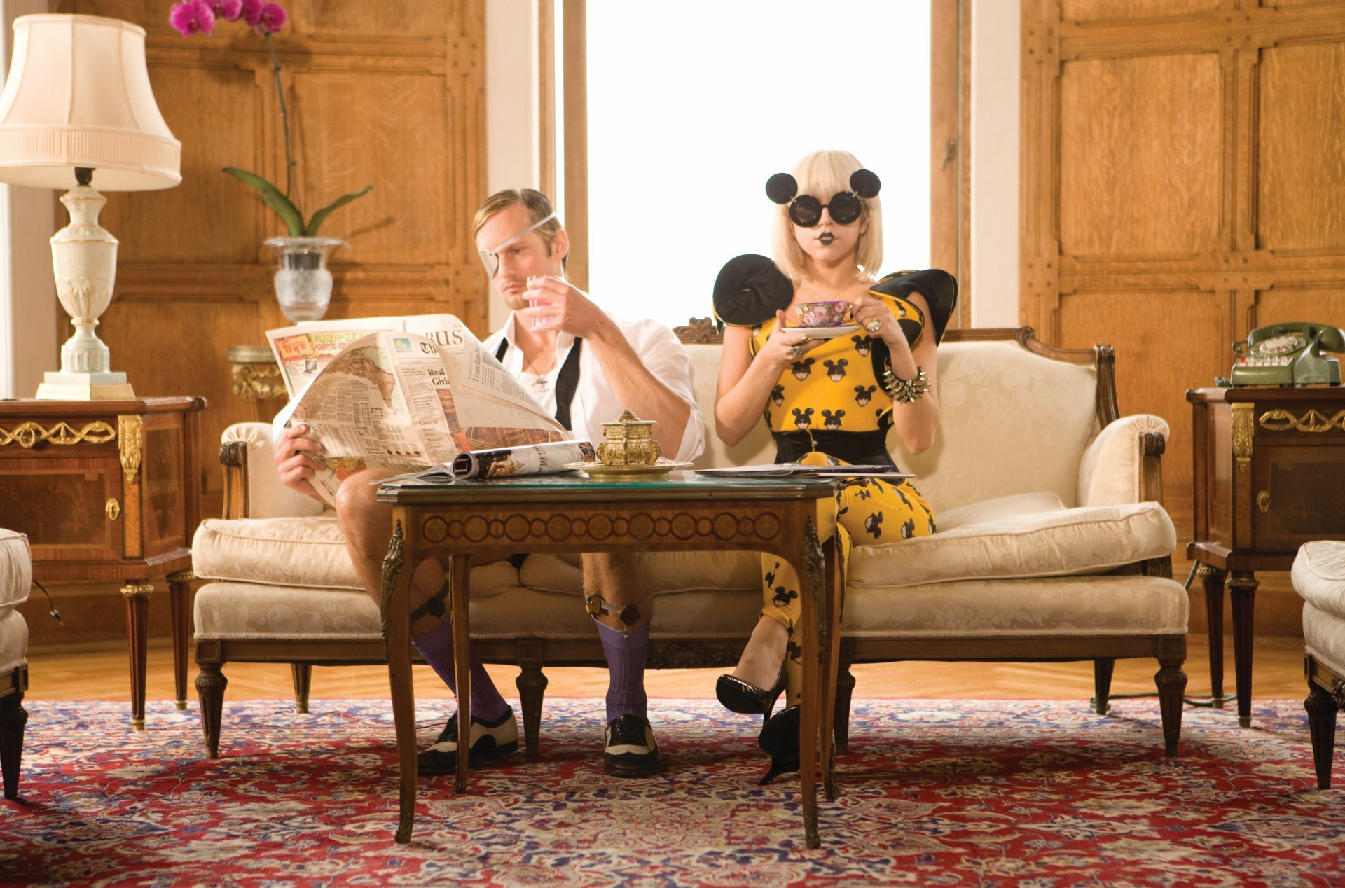 American singer Lady Gaga with blonde hair and sunglasses sits on a sofa next to a man reading a newspaper, with a cup on the table and both wearing socks in an elegant room.