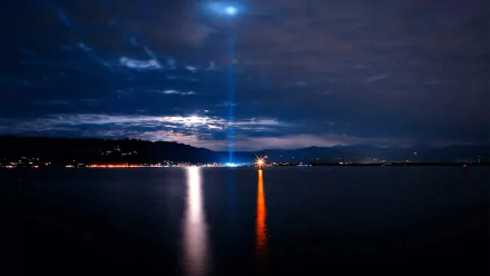 Night photography: calm sea reflecting a blue searchlight and orange beacon under a clouded sky, HD PC desktop wallpaper background.