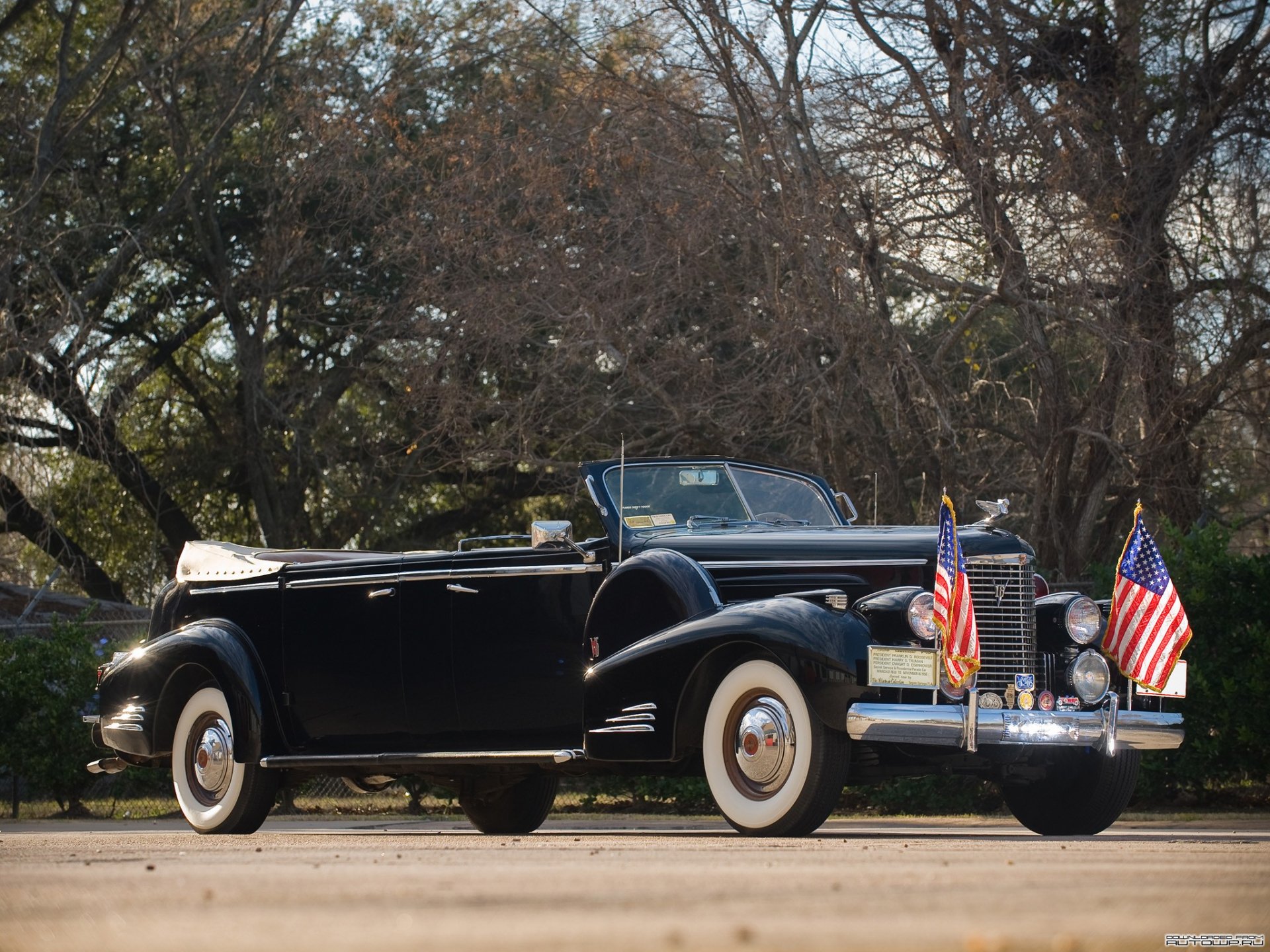 HD PC desktop wallpaper of a black 1938 Cadillac V16 convertible vehicle with whitewall tires and twin American flags, set against a tree-lined background.
