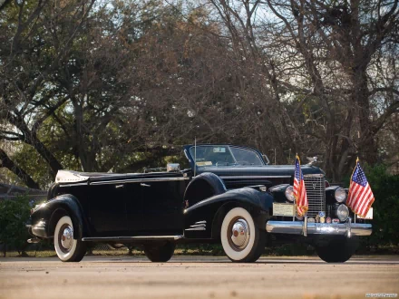 HD PC desktop wallpaper of a black 1938 Cadillac V16 convertible vehicle with whitewall tires and twin American flags, set against a tree-lined background.
