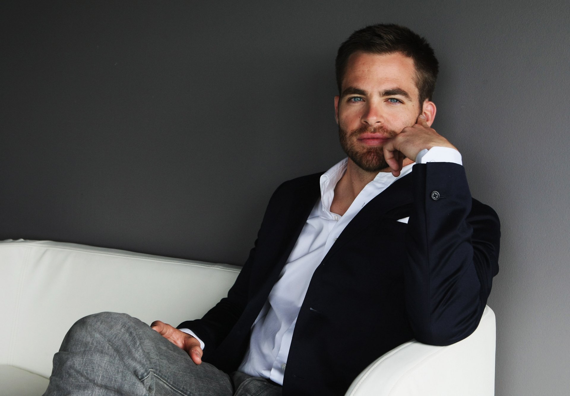 HD PC desktop wallpaper featuring celebrity Chris Pine seated on a white couch against a dark background, dressed in a navy blazer and white shirt.