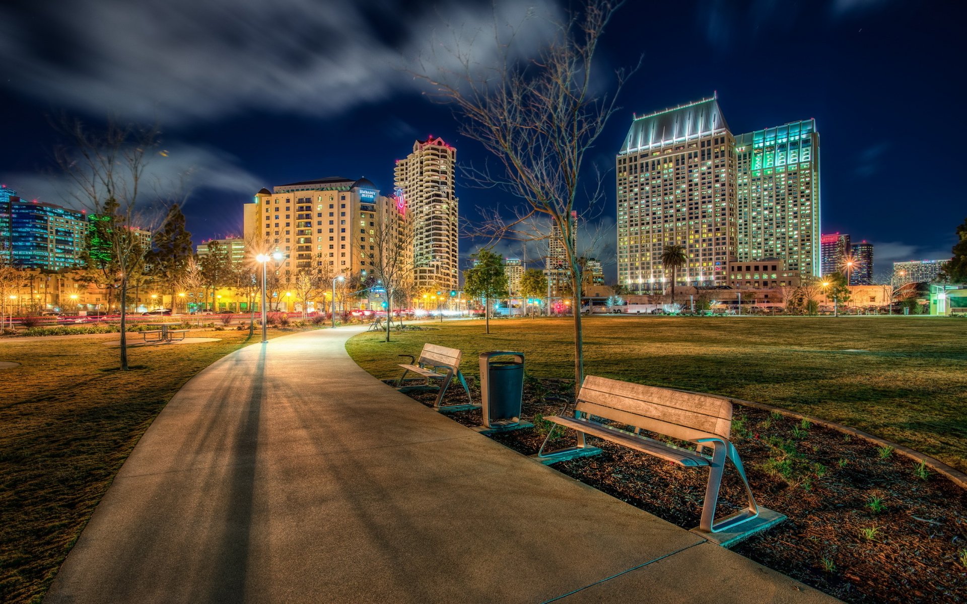 HD PC desktop wallpaper showing a man-made San Diego cityscape at night with illuminated buildings, park benches, and a winding pathway under a cloudy sky.