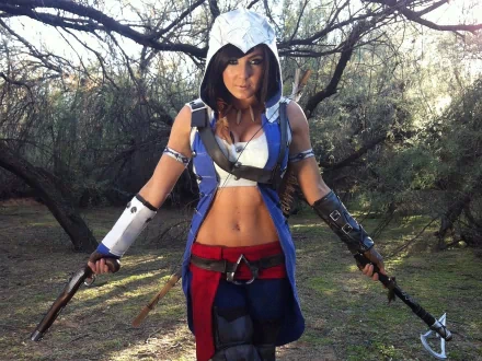 HD desktop wallpaper featuring a woman in detailed cosplay, set against a natural forest background. She wears a hooded costume and holds weapons while standing among trees.