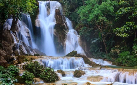 A vibrant HD desktop wallpaper featuring the lush tropical forest and cascading waters of Kuang Si Waterfall in Laos.