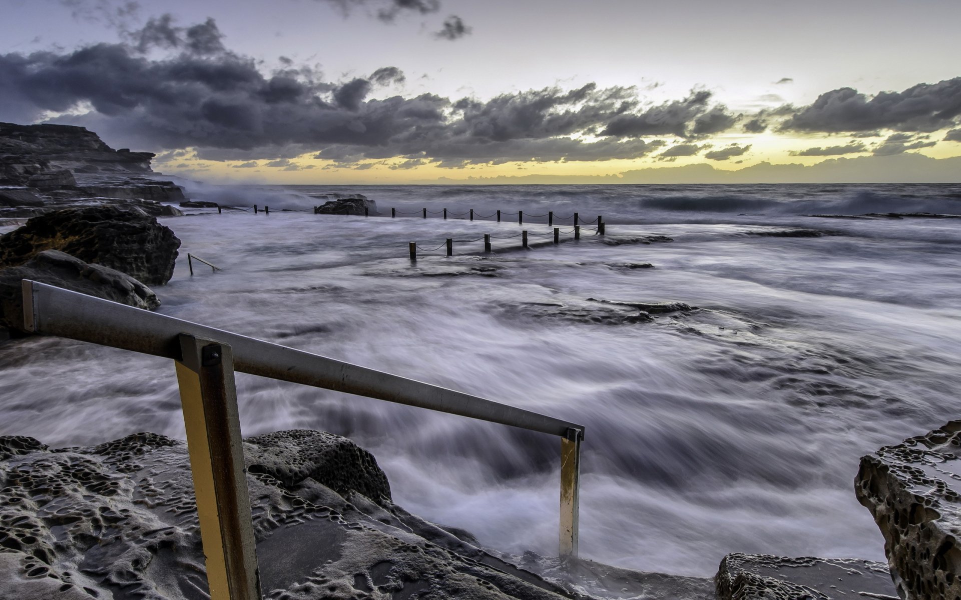 Photography ocean seascape: moody sunrise, waves rushing over rocky shore with a metal handrail and tidal pool fence — 2K Quad HD PC desktop wallpaper/background.