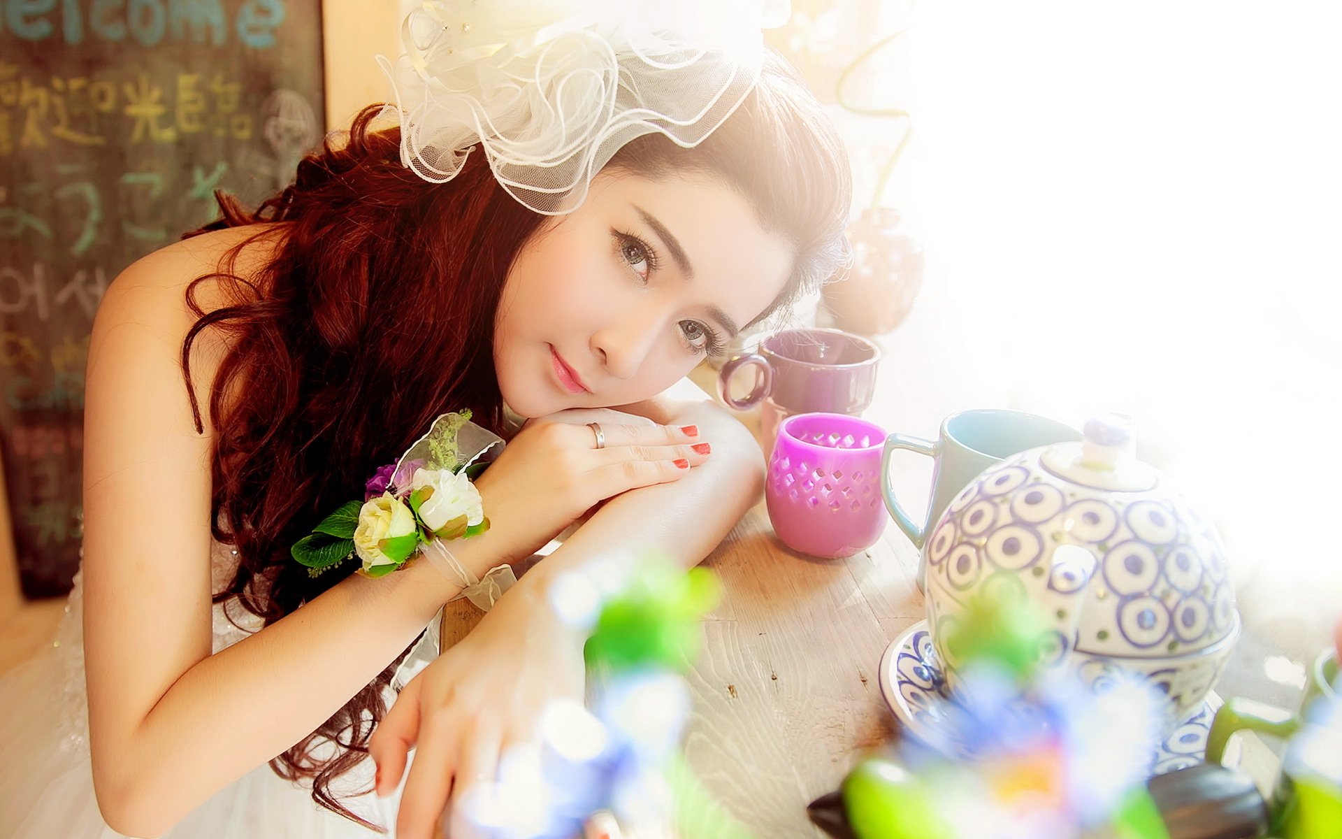A serene Asian woman rests her chin on her hand, surrounded by colorful cups and a decorative teapot, illuminated by soft light in this HD desktop wallpaper.