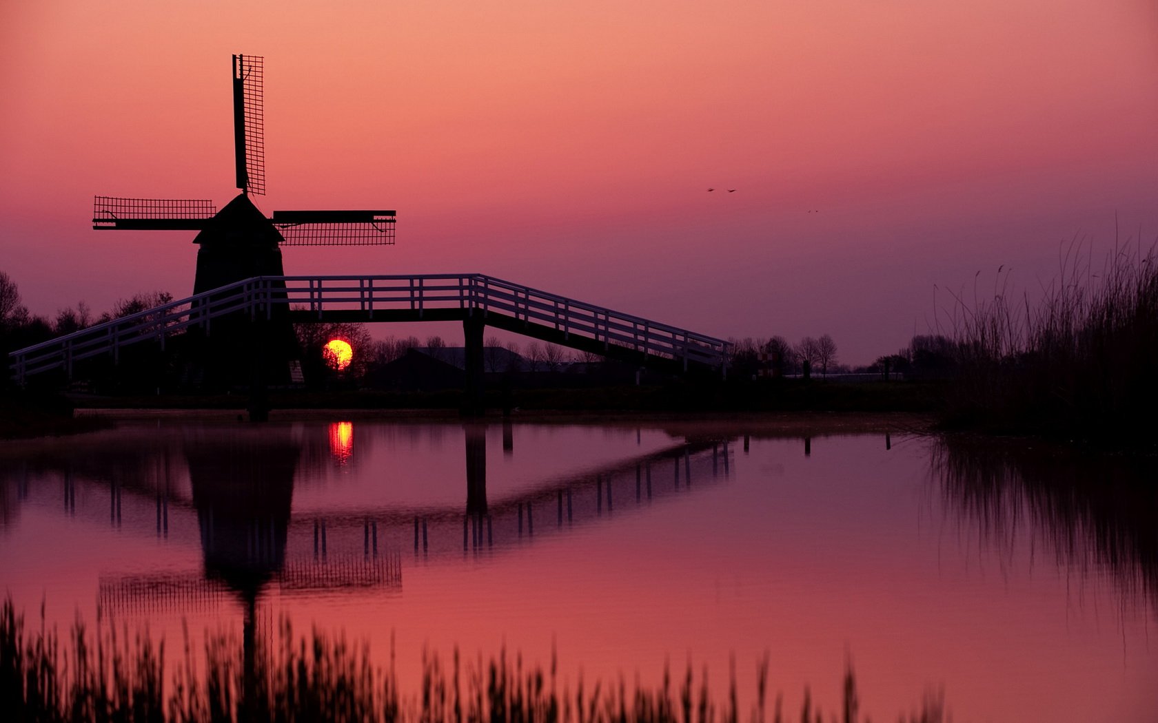 HD desktop wallpaper featuring a man-made windmill silhouetted against a vibrant sunset, reflected in calm water with soft purple and orange hues.