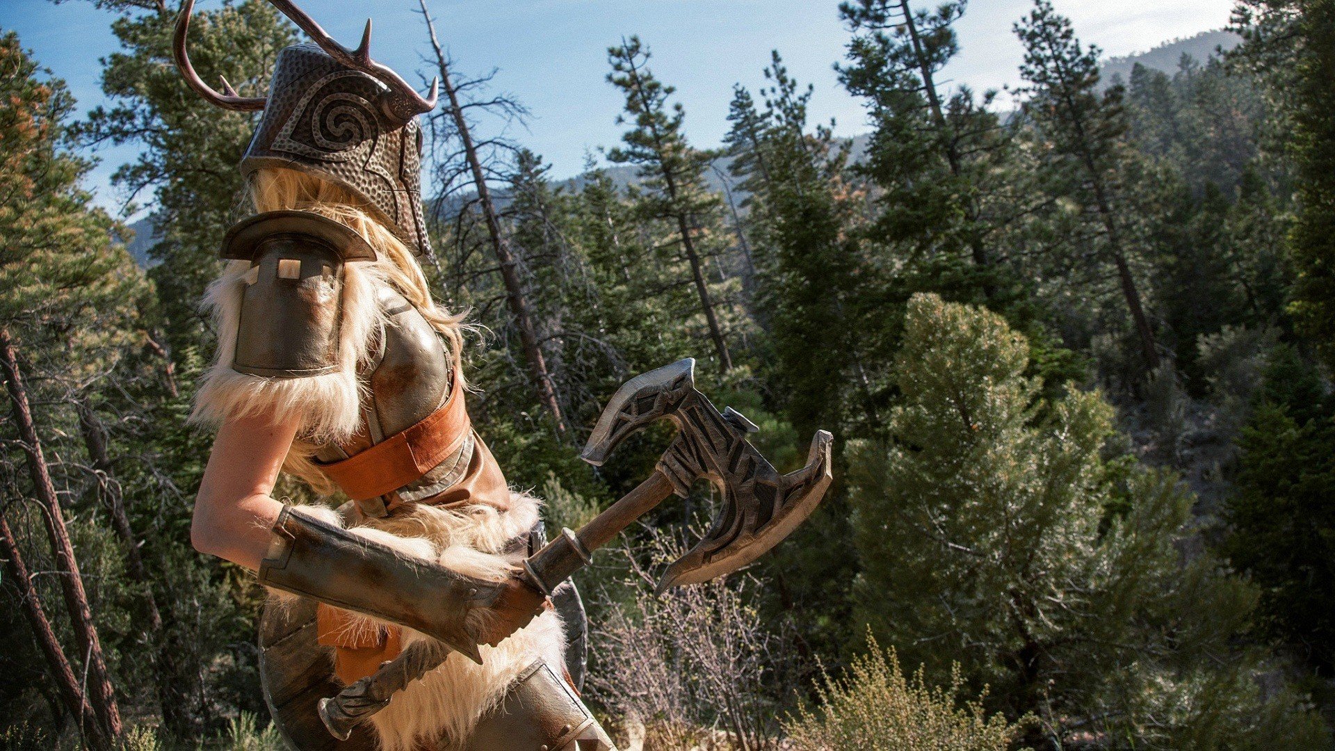 A woman in detailed cosplay stands in a forest, wearing a horned helmet and wielding an axe, surrounded by tall trees. This image serves as a striking HD desktop wallpaper.