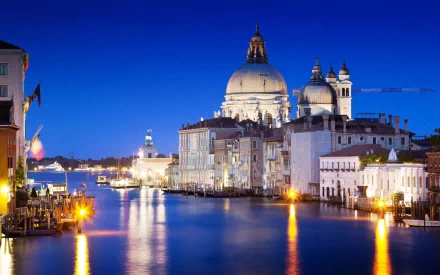 HD desktop wallpaper of Venice at dusk, showcasing iconic man-made architecture along the canal with illuminated buildings reflecting on the calm water under a deep blue sky.
