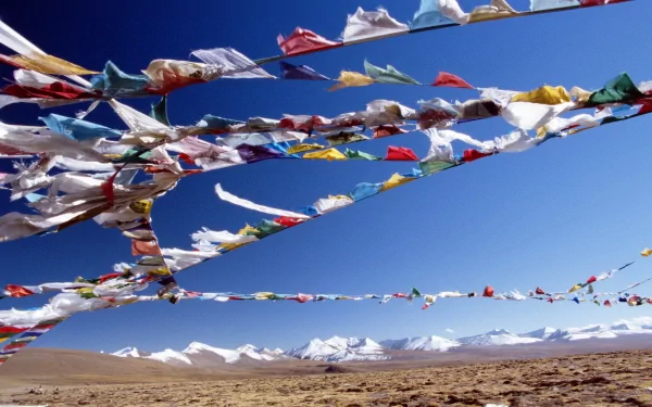 Man-made colorful Tibetan prayer flags sweeping across a remote Himalayan plateau in Nepal — vivid HD PC desktop wallpaper background with snow-capped peaks beneath a clear blue sky.