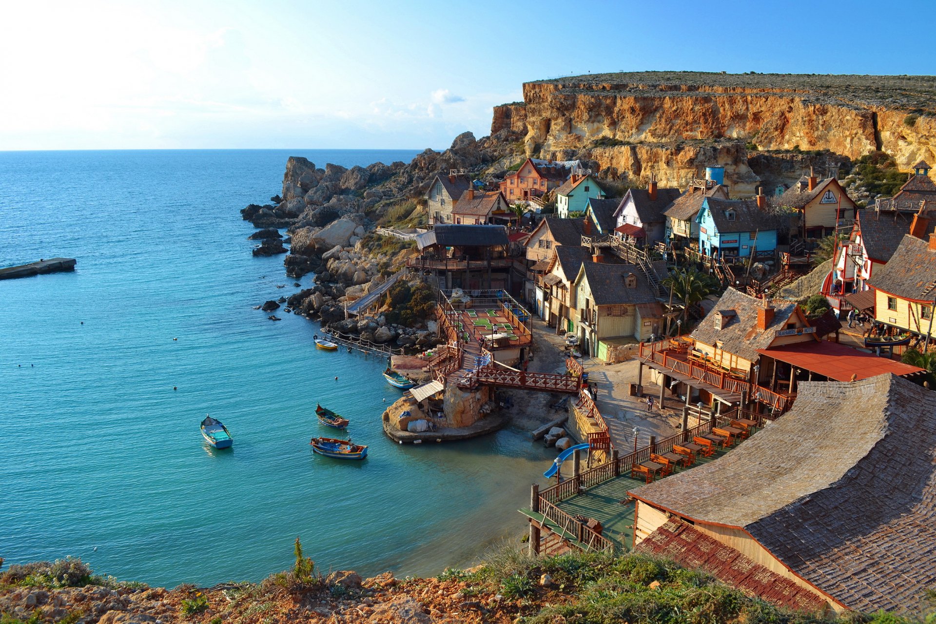 Scenic HD desktop wallpaper featuring the colorful man-made Popeye Village nestled along a rocky coastline with calm blue waters under a clear sky.