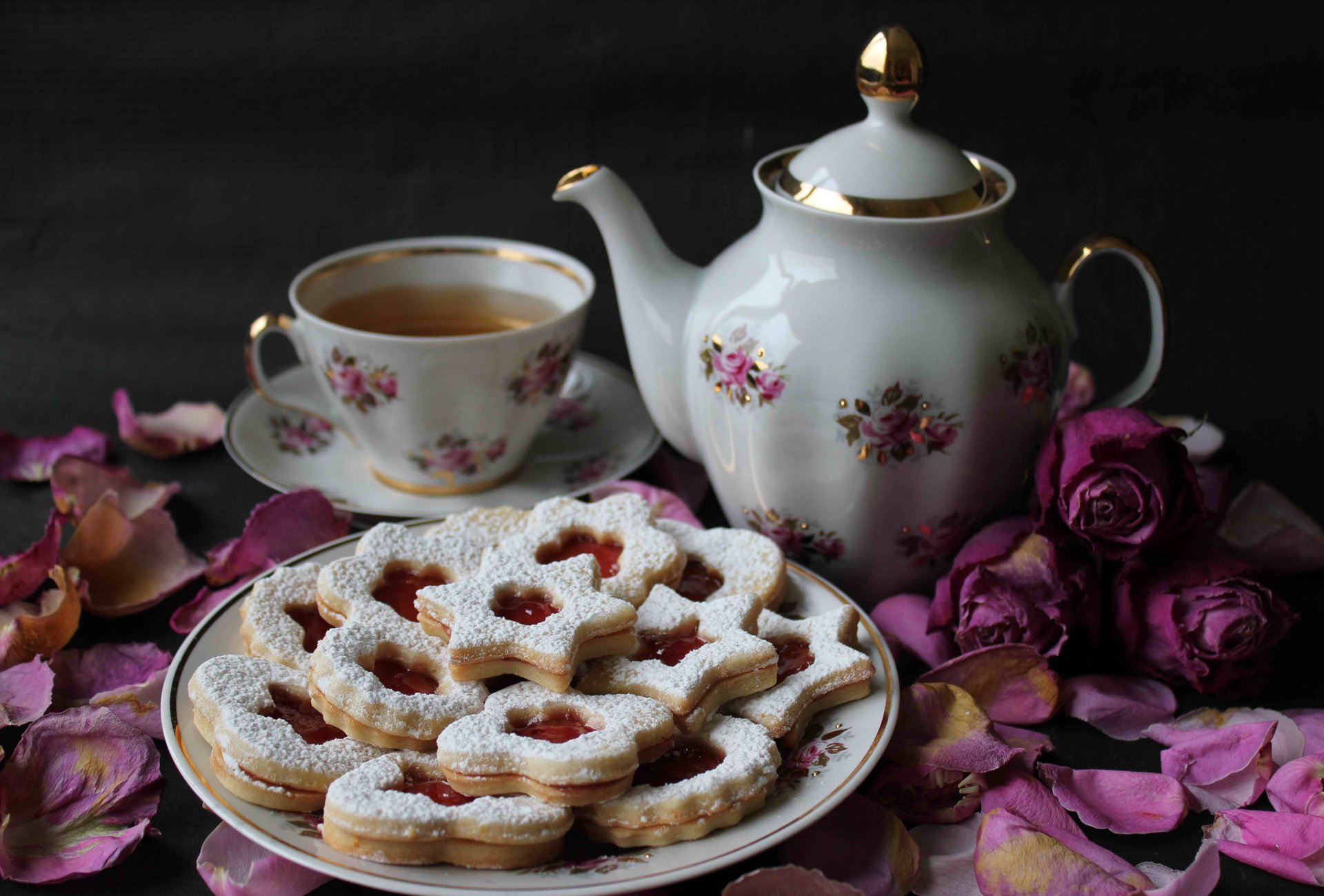 HD PC desktop wallpaper featuring a plate of powdered sugar-dusted cookies with jam centers, a floral teapot, matching teacup, and scattered rose petals.