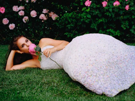 HD PC desktop wallpaper featuring celebrity Natalie Portman lying on grass in a white gown, holding a pink rose with a backdrop of blooming roses and greenery.