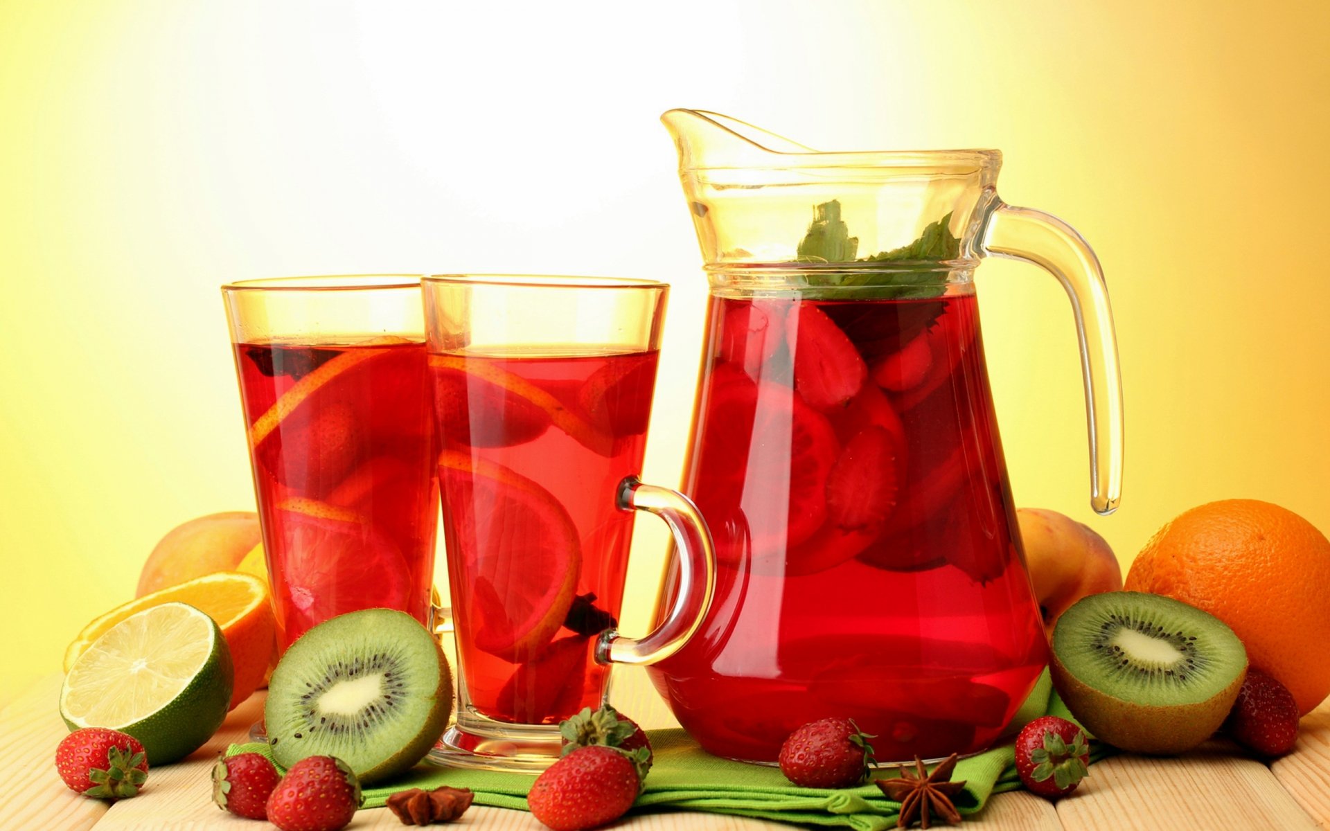 A vibrant pitcher of fruit punch sits beside two glasses filled with the refreshing drink, surrounded by fresh strawberries, kiwi slices, and citrus fruits, all against a warm yellow background.