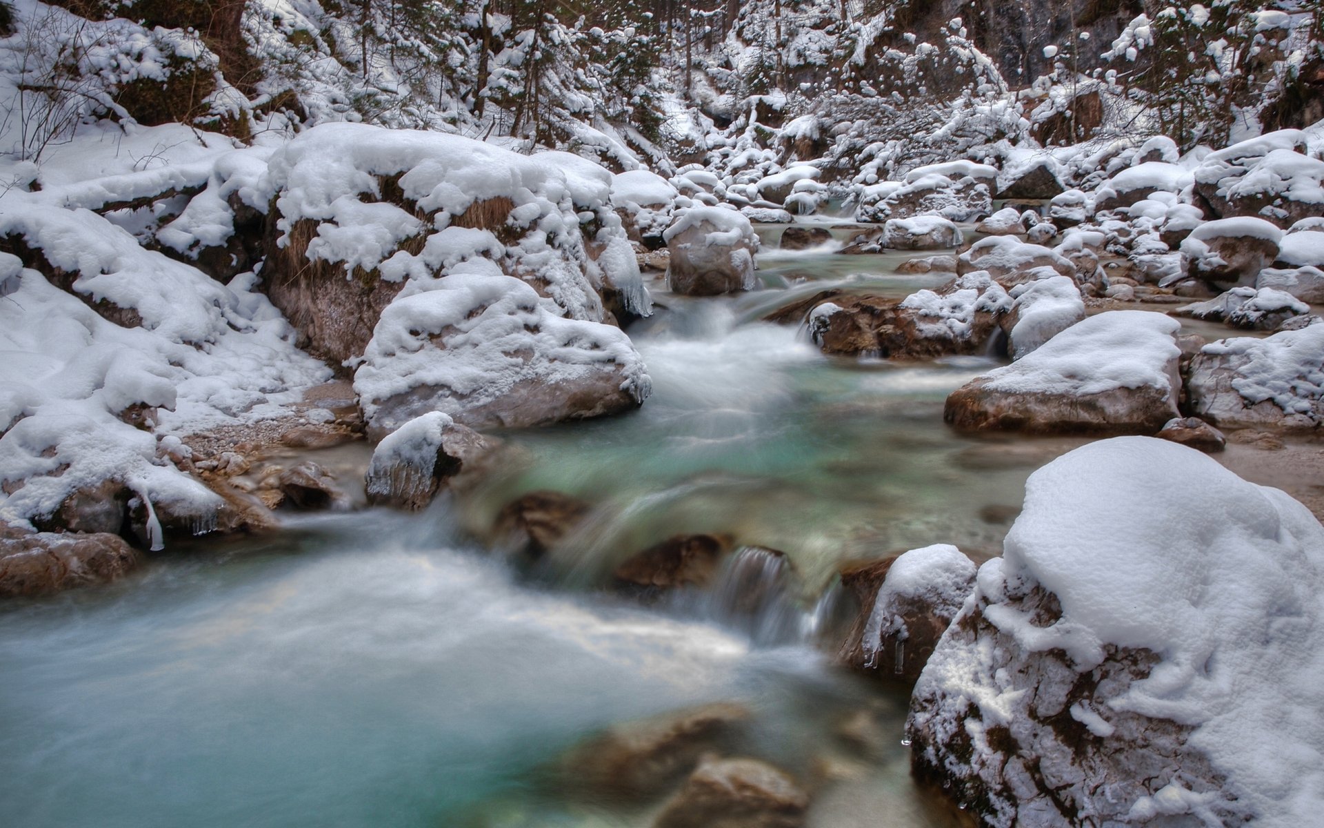 HD desktop wallpaper of a serene winter stream flowing through a snow-covered forest, showcasing tranquil nature in vivid detail.