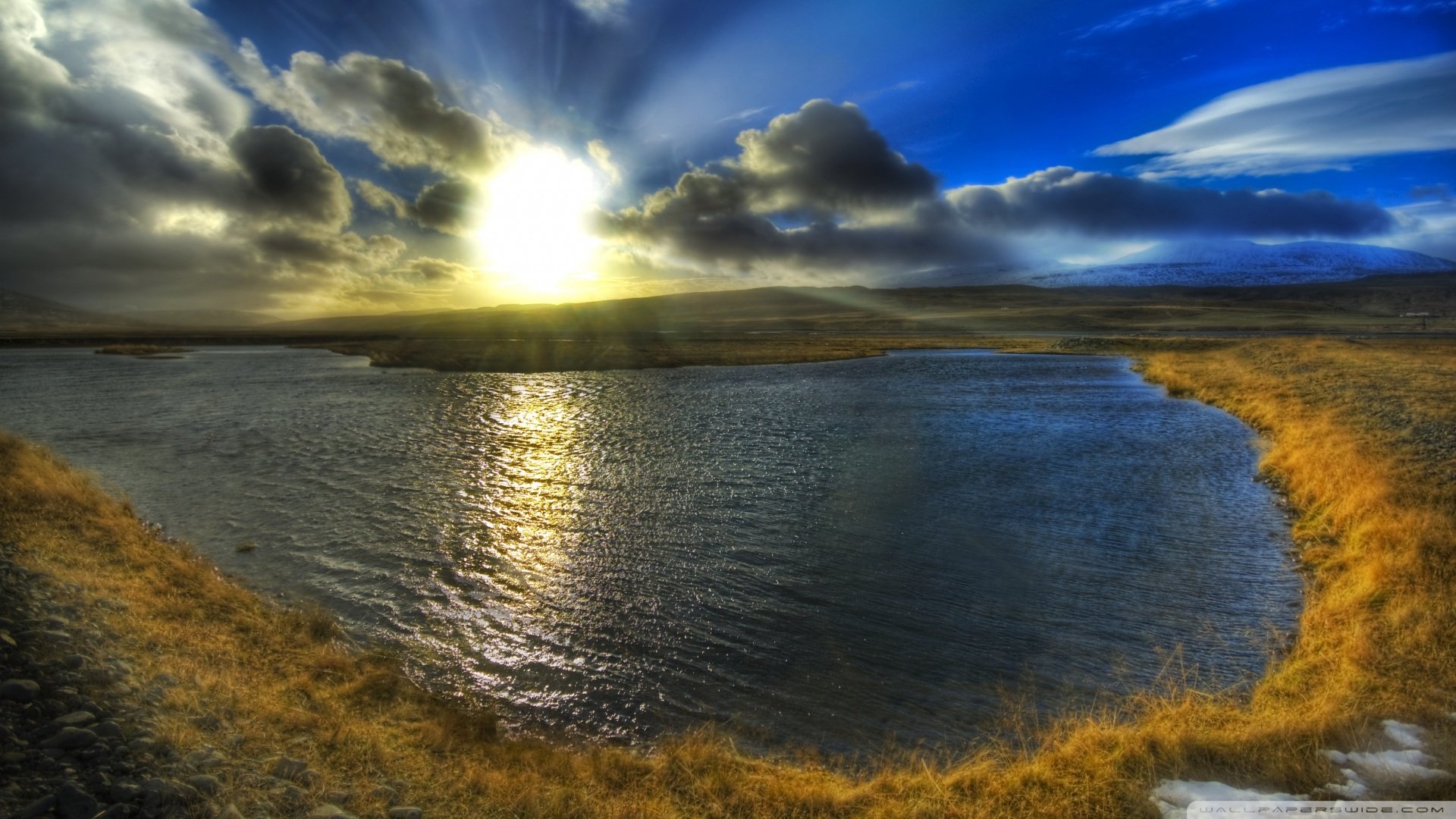 A stunning Icelandic landscape featuring a serene lake reflecting a sunbeam, surrounded by grass and dramatic clouds under a vibrant sky. Captured in HDR for enhanced detail.
