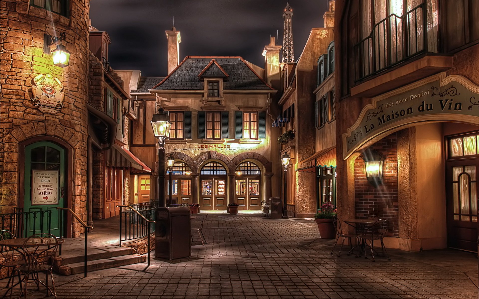 Charming nighttime view of a French-inspired street, featuring man-made buildings with warm light illuminating the cobblestone path, creating an inviting atmosphere. HD desktop wallpaper.