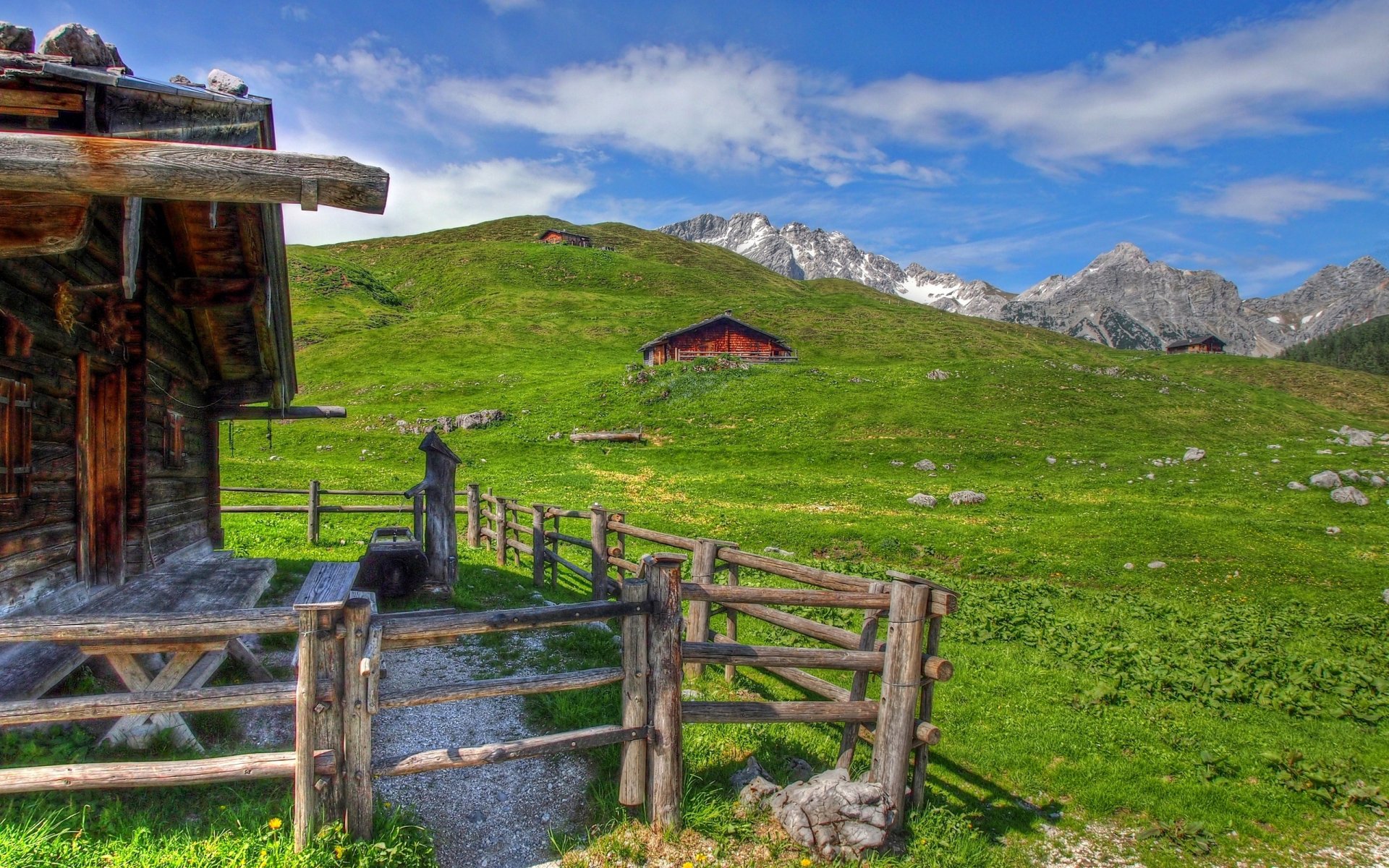 HD desktop wallpaper of a man-made wooden cabin nestled in a lush green hillside with a mountain range under a clear blue sky.