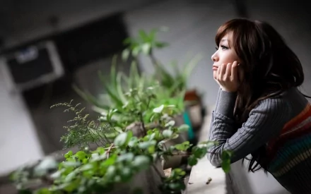 An Asian woman gazes thoughtfully in the distance while leaning on a surface with lush green plants. The image serves as an HD desktop wallpaper and background.