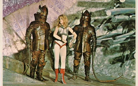 Barbarella movie scene: heroine in white jumpsuit and red boots flanked by two armored sentinels in a retro sci‑fi set — 2K Quad HD PC desktop wallpaper background