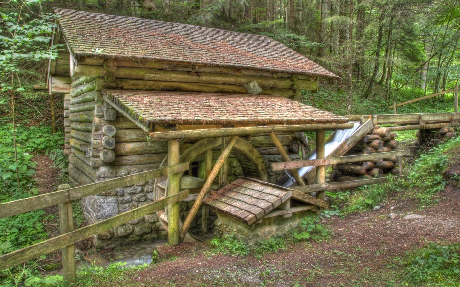 HD desktop wallpaper of a man-made watermill nestled in a lush forest, showcasing rustic wooden logs and a flowing water wheel.