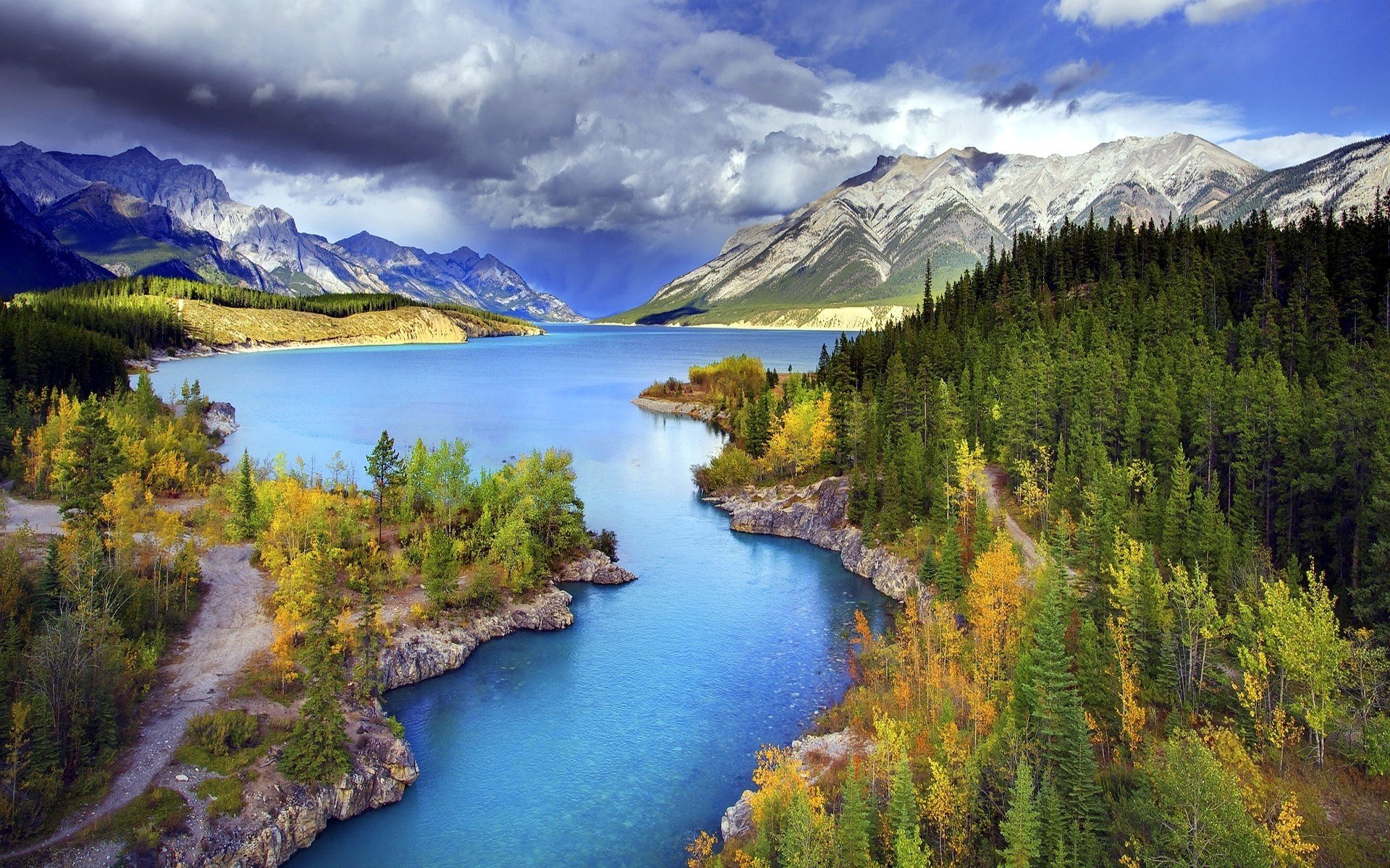A breathtaking landscape featuring mountains under a dramatic sky, lush forests, and a serene river, showcasing the vibrant colors of fall in nature. A stunning HD desktop wallpaper.
