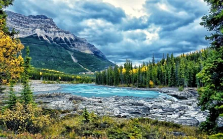 Stunning view of Athabasca Falls, showcasing a vibrant landscape of turquoise waters, rocky terrain, and lush greenery framed by dramatic mountains and a moody sky.