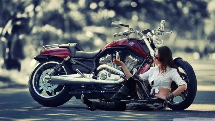 A woman in a white shirt and leather pants sits on the ground, leaning against a red Harley-Davidson motorcycle on a blurred background. This HD wallpaper captures the spirit of girls and motorcycles.