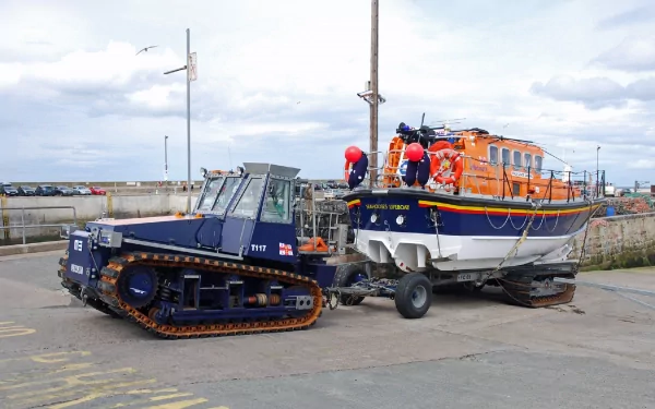 A powerful tractor and an orange rescue boat are parked at the harbor, showcasing nautical equipment against a cloudy sky. This HD wallpaper highlights the strength of marine vehicles.