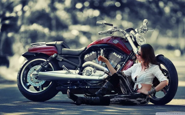 A woman in a white shirt and leather pants sits on the ground, leaning against a red Harley-Davidson motorcycle on a blurred background. This HD wallpaper captures the spirit of girls and motorcycles.