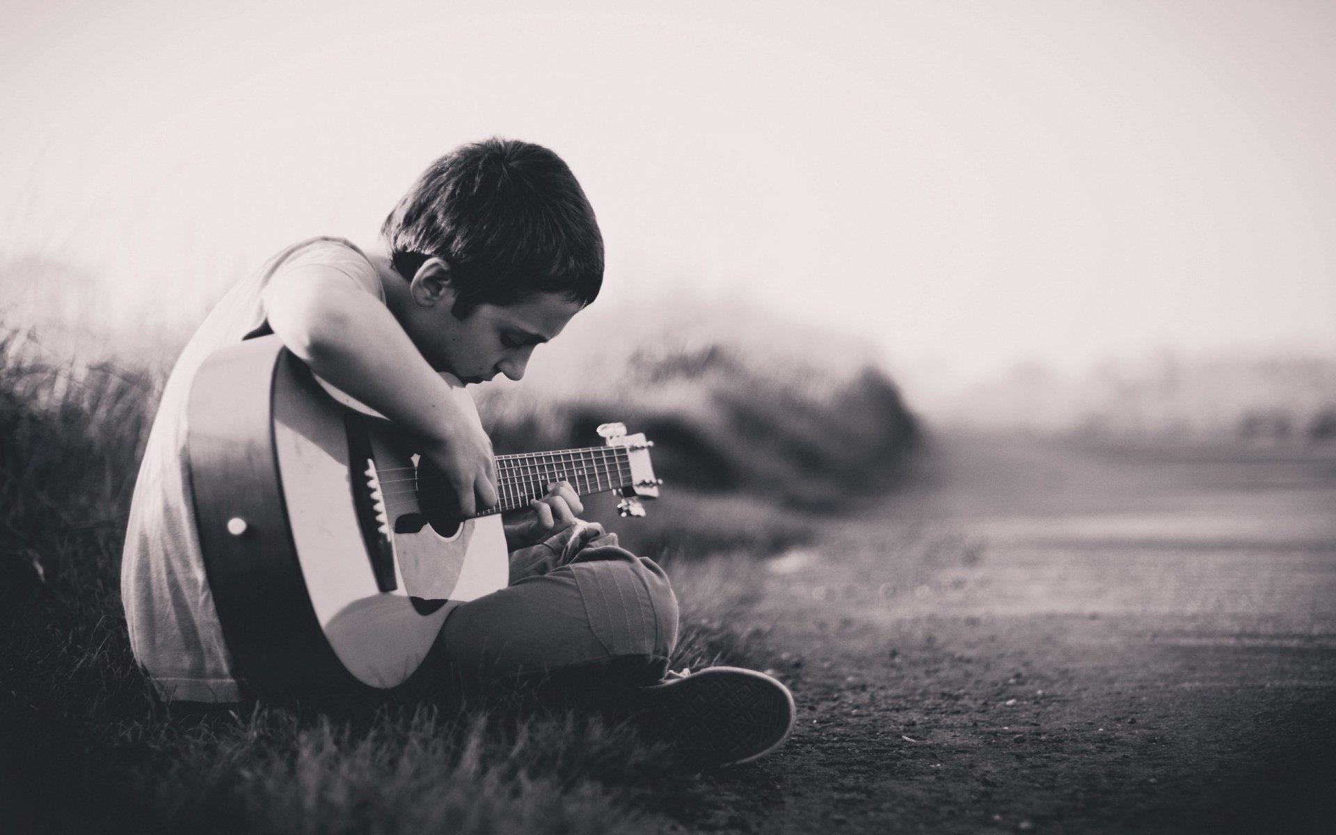 Black and white HD desktop wallpaper of a contemplative young man sitting outdoors, playing an acoustic guitar, capturing a reflective mood.