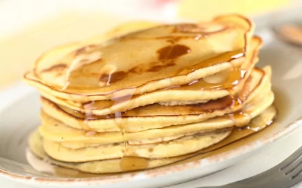 2K Quad HD PC desktop wallpaper/background showing a close-up stack of golden pancakes drizzled with syrup on a white plate, warm breakfast food shot