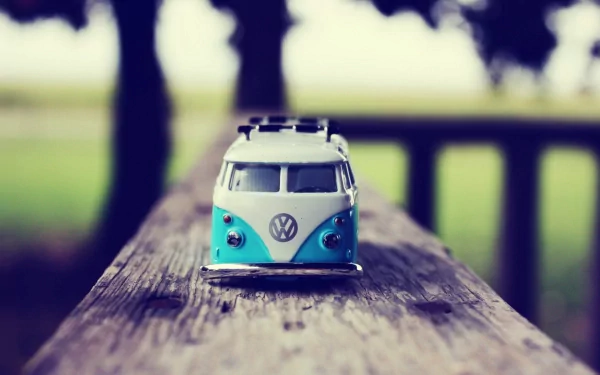 A close-up of a toy Volkswagen van resting on a weathered wooden surface, set against a blurred outdoor background, creating a nostalgic and vibrant scene.