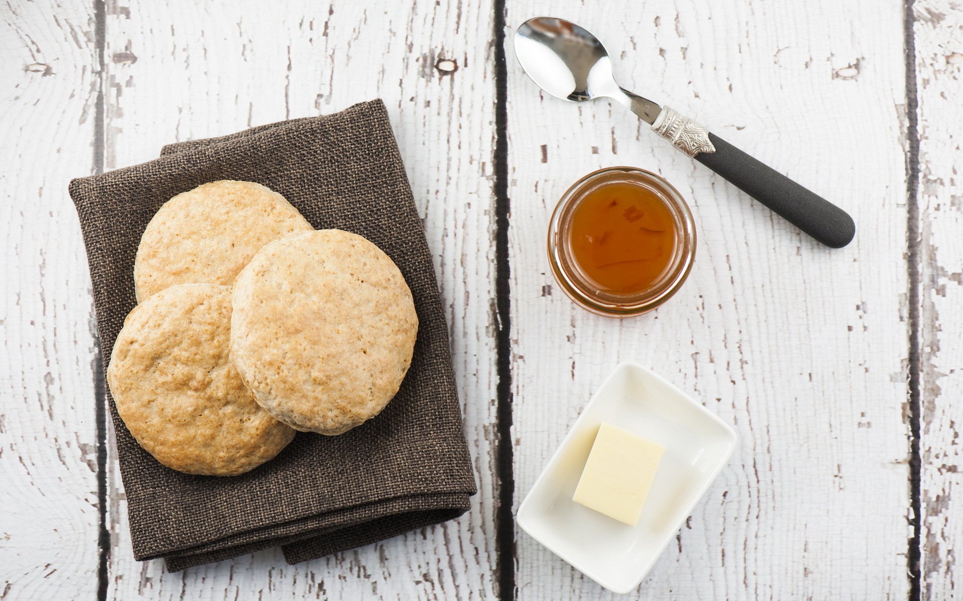 A cozy arrangement of freshly baked cookies on a brown napkin, accompanied by a dish of butter and a jar of honey, set against a rustic wooden background.