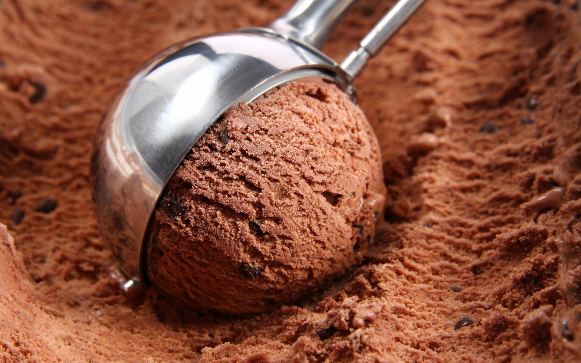 Close-up of a metal ice cream scoop resting on rich, chocolate ice cream, set against a textured background, creating a delicious desktop wallpaper.