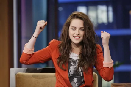 HD desktop wallpaper and background featuring celebrity Kristen Stewart, smiling and raising her arms, dressed in a casual red jacket and printed T-shirt, set in a modern indoor setting.