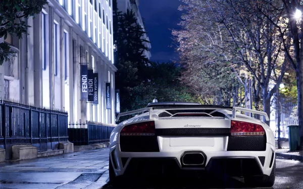 HD desktop wallpaper featuring a white Lamborghini Murcielago parked on a quiet, tree-lined city street at night. The sleek design and rear view of the car are prominently displayed.