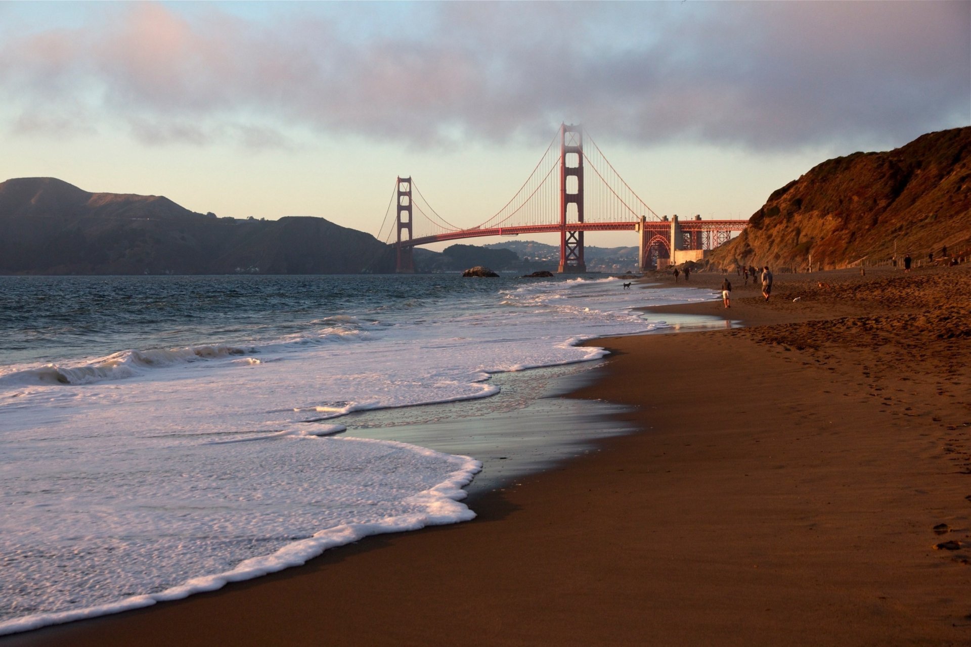 HD desktop wallpaper featuring the man-made Golden Gate Bridge viewed from a sandy beach at sunset with waves gently washing ashore.