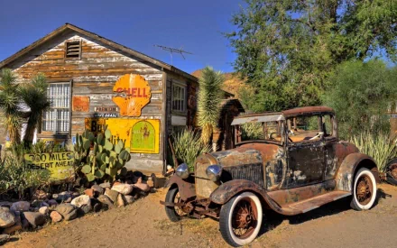 A vintage scene featuring a weathered car parked near a rustic wooden building adorned with colorful Shell advertisements, set against a clear blue sky and desert landscape.