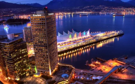 A stunning HD image of Vancouver at night, showcasing the city’s skyline, waterfront with colorful lights, and majestic mountains in the background. A captivating man-made landscape.